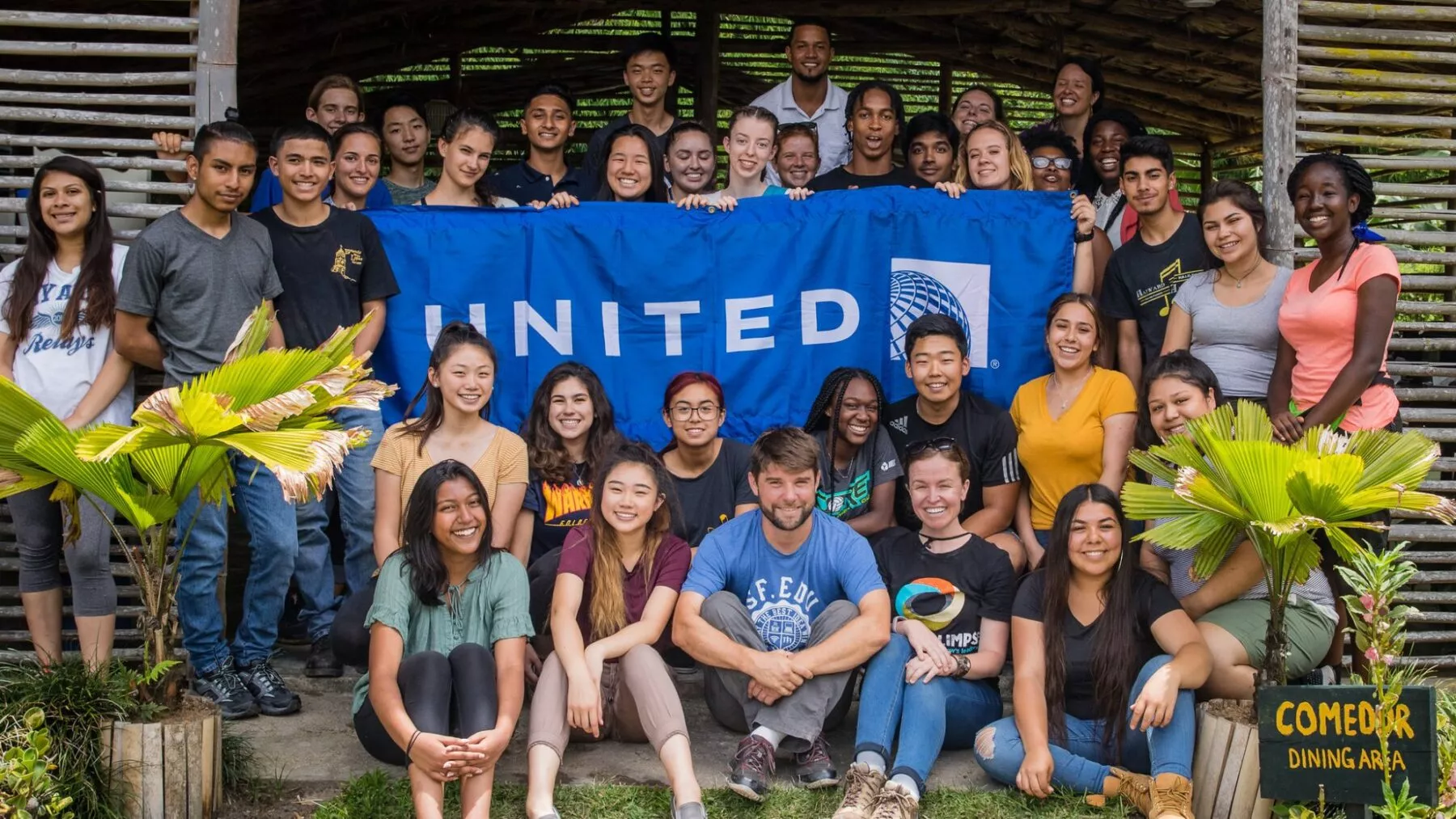 Group of Global Glimpsers stand and sit around a United Airlines flag being held up. Looking at the camera