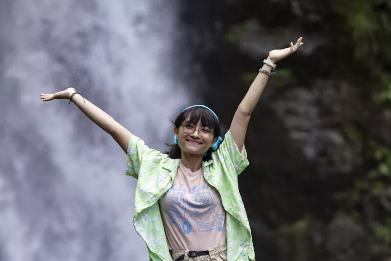 Picture of a girl standing in front of a waterfall and posing for the camera with her arms up in the air